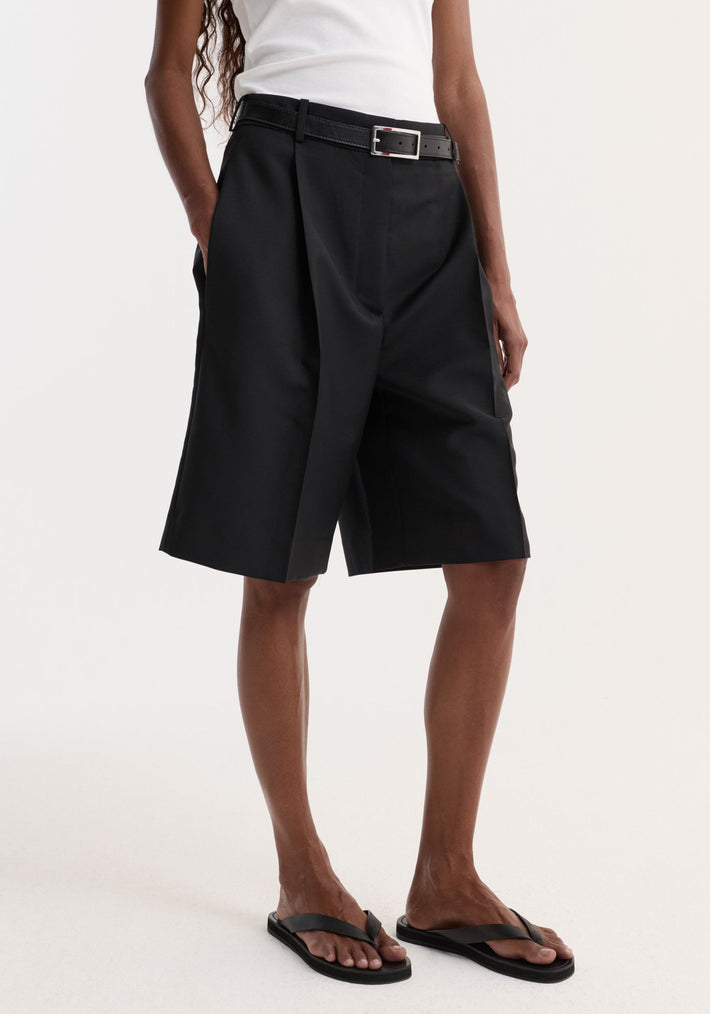 Tailored single pleat shorts in black, featuring knee-length design, side pockets, and a slim black belt, highlighted against a light background.