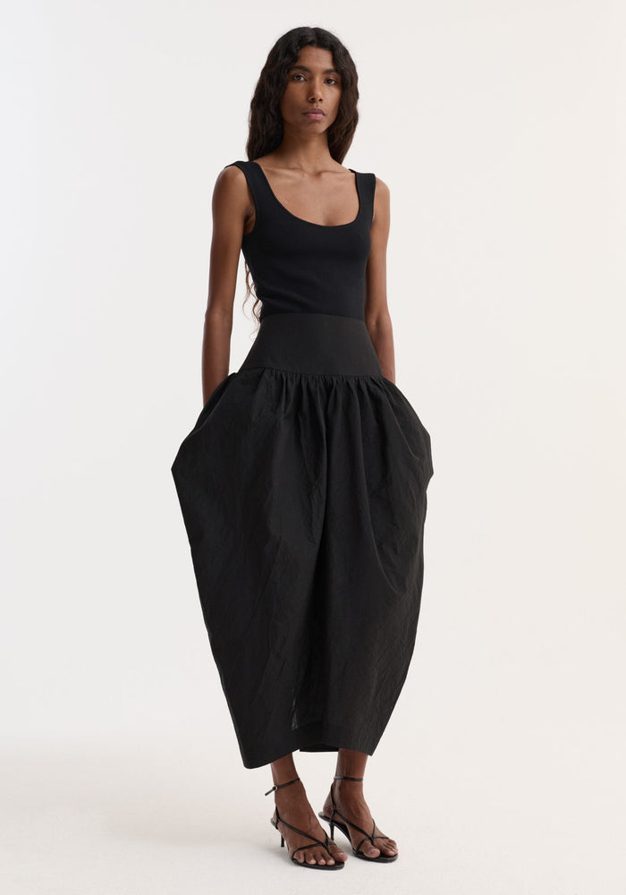 Model wearing the Sculptural volume skirt | black, featuring a mid-waist, fitted waistband, and architectural pockets, paired with a sleeveless top and strappy heeled sandals.