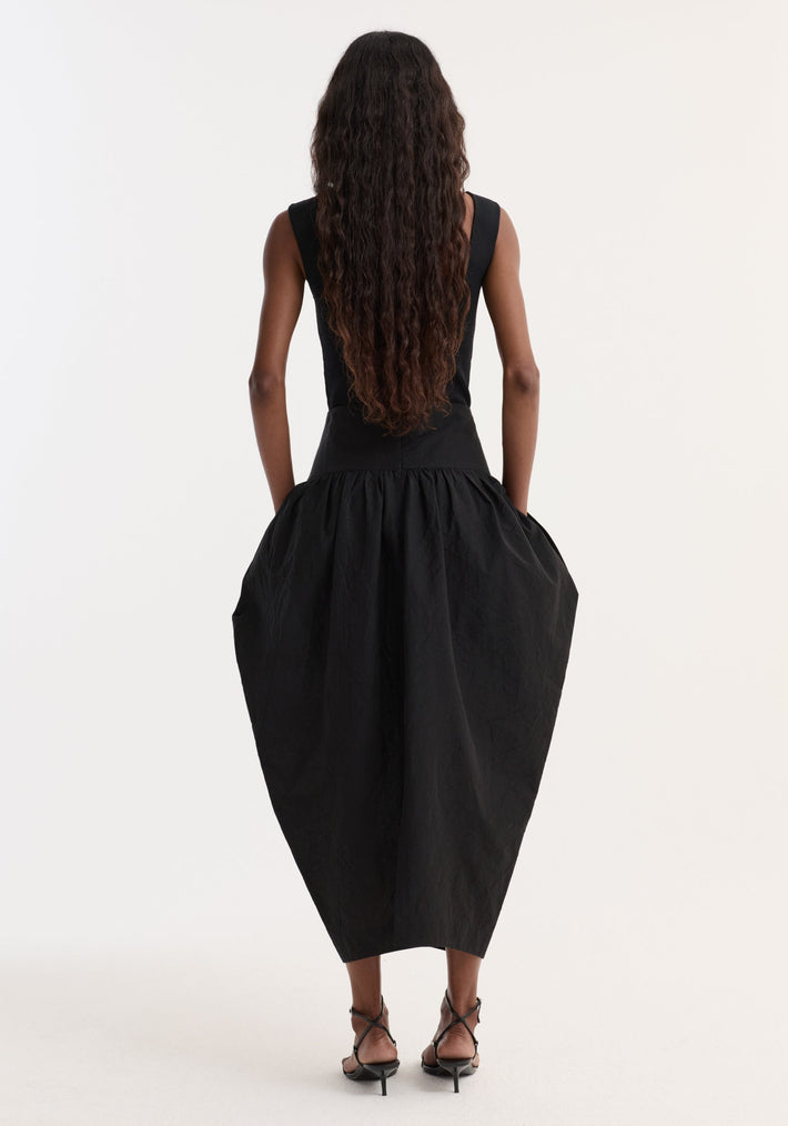 Back view of a model wearing a Sculptural volume skirt with a fitted waistband and architectural pockets, creating a trapezium silhouette, paired with strappy black heeled sandals.
