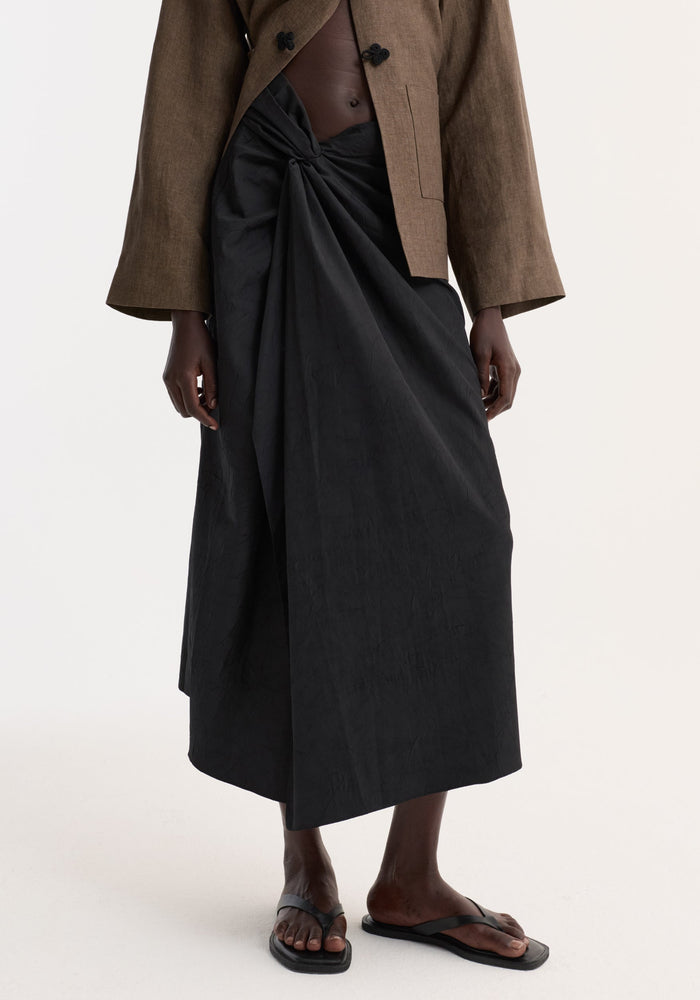 Model wearing Sarong textured skirt | black, featuring a knot detail and gathered fabric at the waist, paired with flat thong sandals.