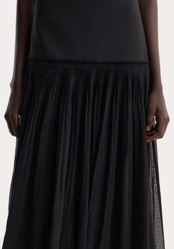 Pleated muslin halter dress features a sleeveless black top with a structured bodice, flowing into a finely pleated, full-length skirt, showcasing elegant movement.