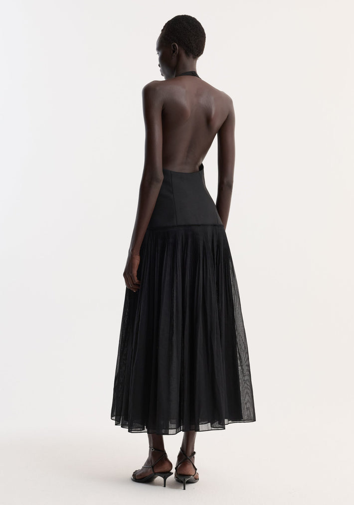 Pleated muslin halter dress | black: Model in backless, fitted bodice dress with pleated mid-calf skirt and strappy heels, viewed from a three-quarter angle.