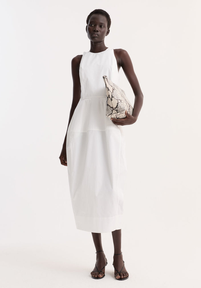 Model wearing Sculptural poplin dress in optic white, featuring a sleeveless design, high neck, trapezium-shaped skirt, and back split; paired with snakeskin pouch and black heels.