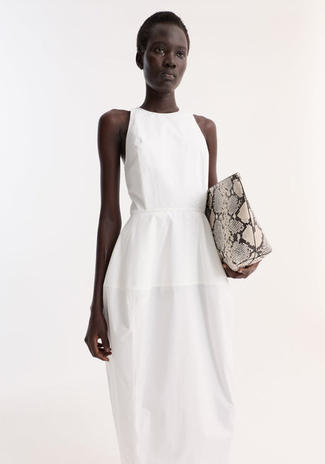 Model wearing Sculptural poplin dress in optic white, featuring a sleeveless design, high neck, trapezium-shaped skirt, and back split; paired with snakeskin pouch and black heels.
