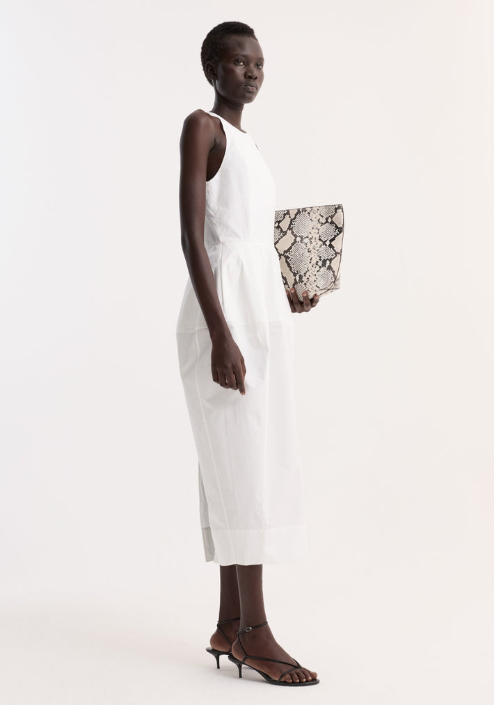 Dark-skinned model in profile wearing the Sculptural poplin dress in optic white, holding a snakeskin clutch, paired with black strappy sandals, showcasing the dress's architectural design.