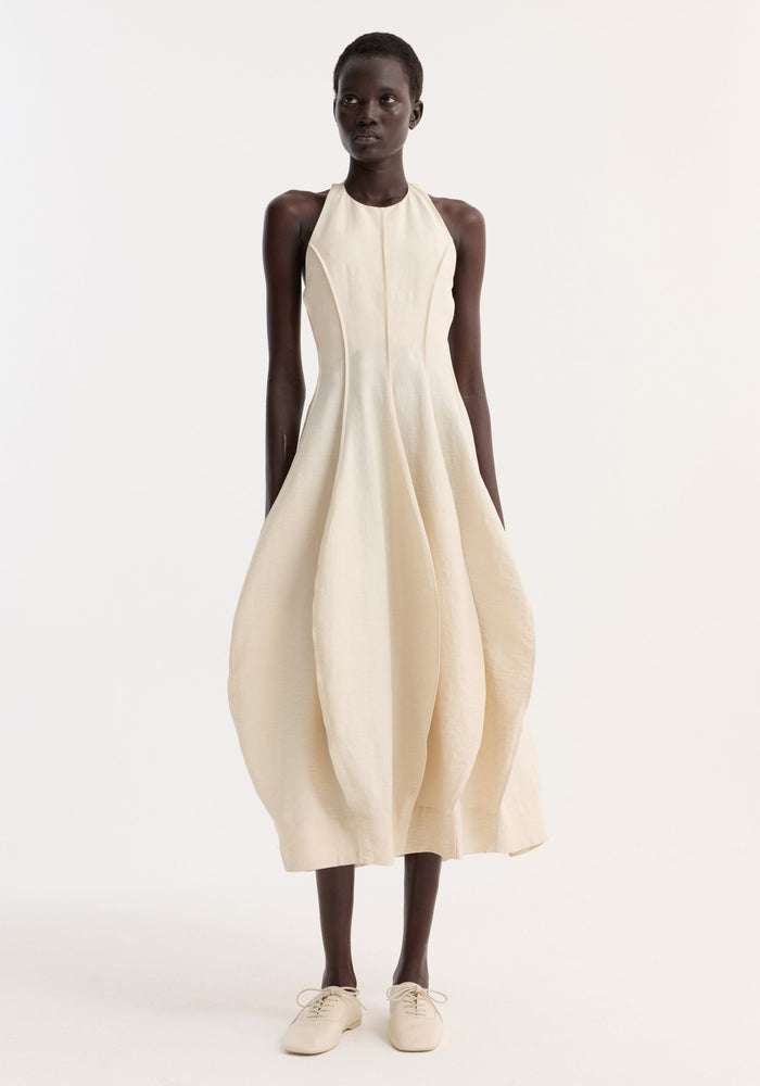 Model wearing the Open Back Tulip Dress | Butter, featuring a halter neck, balloon skirt, and low back, paired with light-beige lace-up flats.