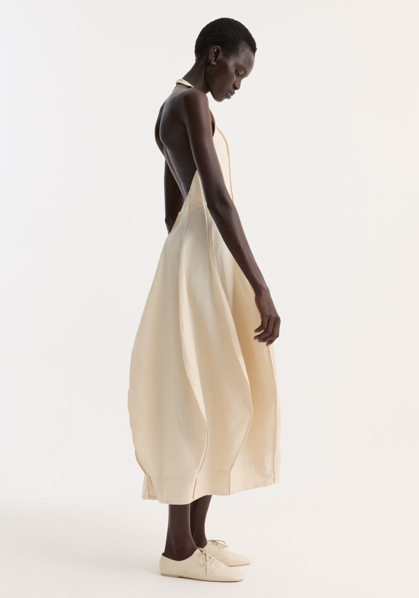 Dark-skinned woman in profile wearing the Open Back Tulip Dress | Butter, showcasing its halter neck, low back, and pleated, balloon-shaped skirt.