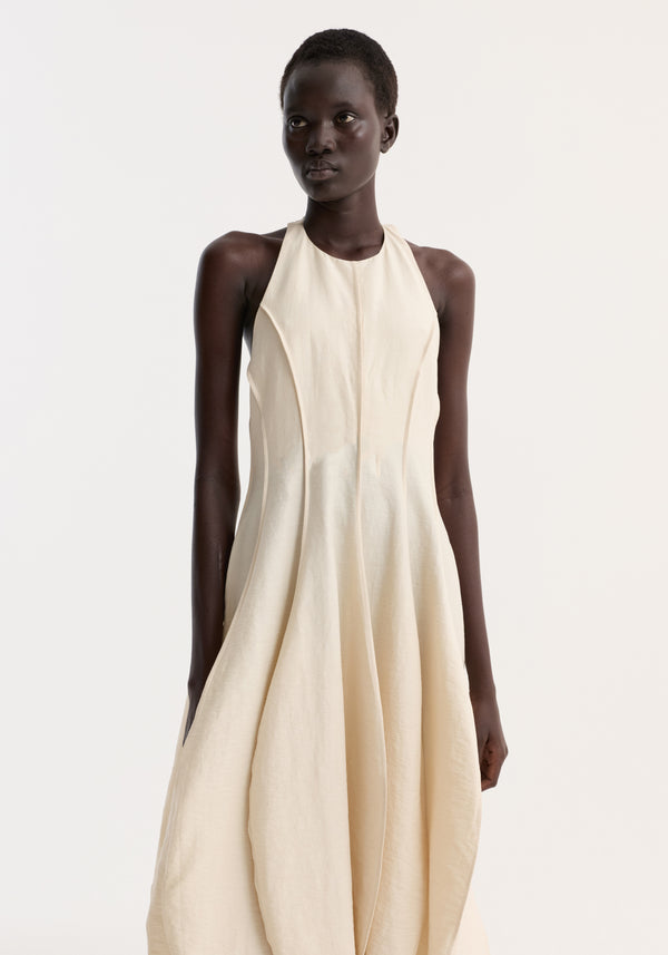Model in a sleeveless Open back tulip dress | butter, showcasing a halter-neck, pleated balloon skirt, high neckline, and low back with a textured finish.