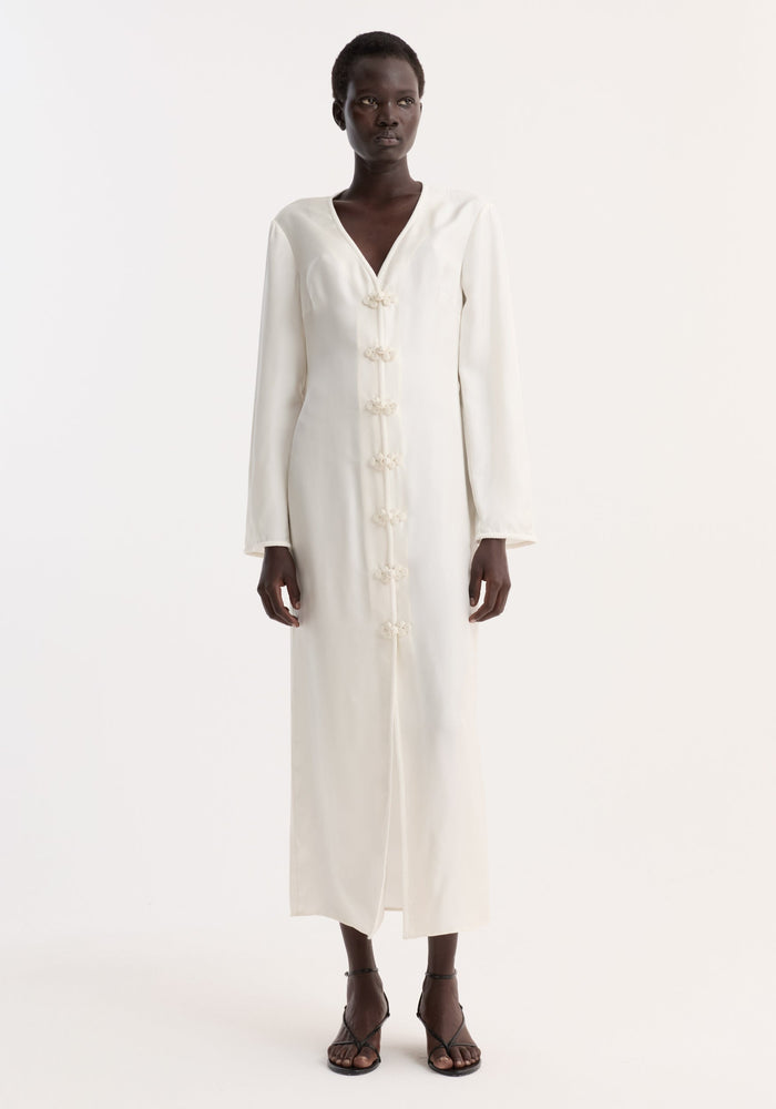Dark-skinned model in a Pankou closure silk dress | cream, featuring frog closures, V-neck, long sleeves, and high front split, styled with black strappy sandals.