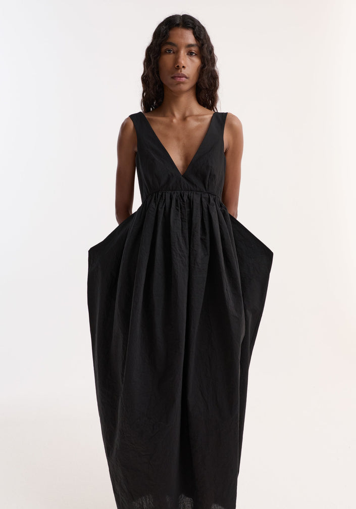 Woman in Sculptural textured cotton dress | black, featuring a deep-V neckline, low back, voluminous skirt, and discreet pockets, paired with black strappy heeled sandals.
