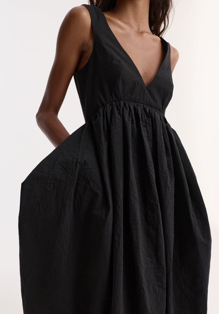 Sculptural textured cotton dress in black: Sleeveless, deep V-neck, gathered voluminous skirt, side pockets, low back, worn by model with hands in pockets.