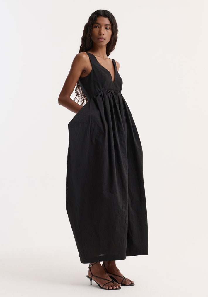 Model in profile wearing the Sculptural textured cotton dress | black, featuring a sleeveless V-neck, gathered waist, trapezium-shaped skirt, and side pockets.