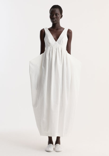 Model in Sculptural textured cotton dress | off white, featuring a sleeveless design, deep V-neckline, full gathered skirt, hidden side pockets, and simple slip-on shoes.