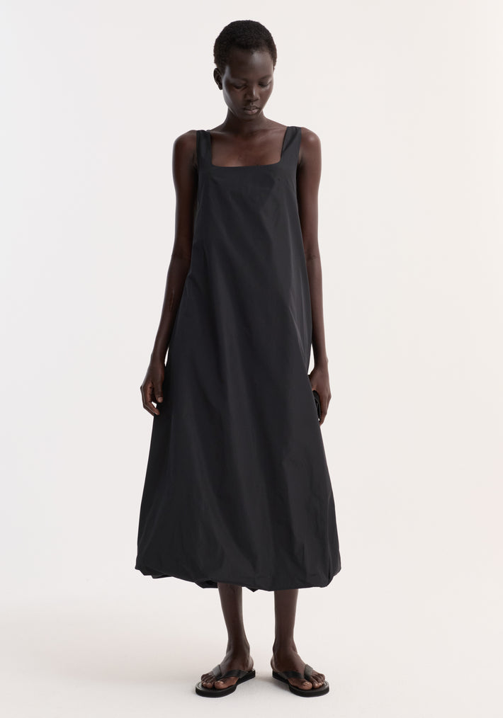 Tall woman in a Technical volume dress | black, featuring wide straps, scooped square neckline, and balloon hemline, paired with black slide sandals.