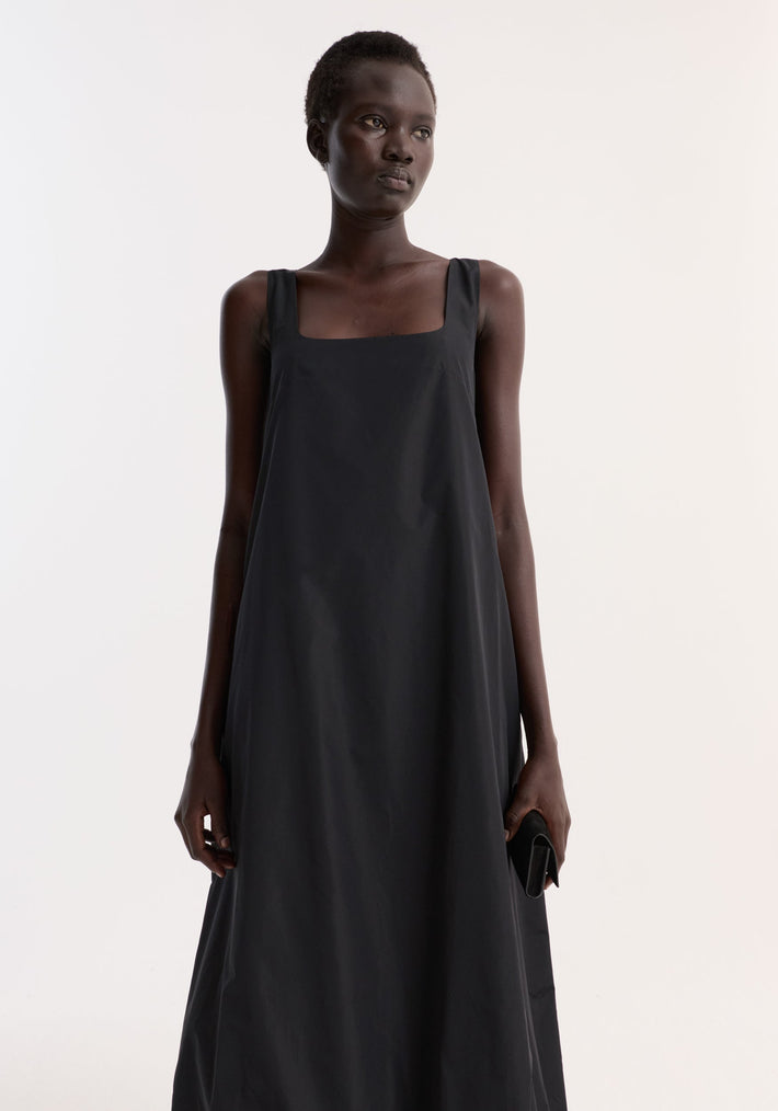 Tall model in Technical volume dress | black, featuring sleeveless design, square neckline, and balloon hemline, holding black clutch against white background.