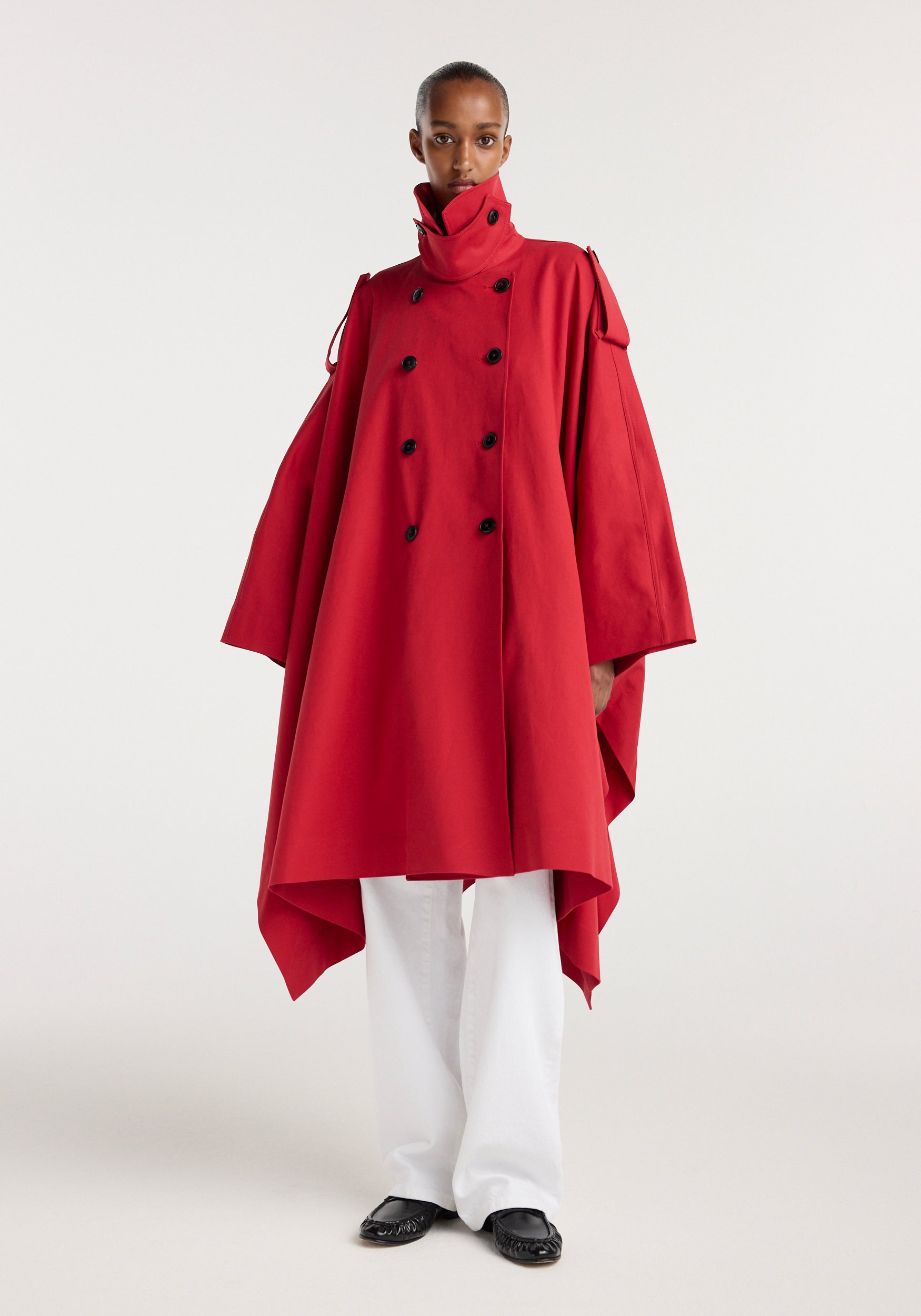 Model wearing a Cape trenchcoat | cherry in vibrant red with black buttons, high collar, and an asymmetrical hem over white pants and black shoes.