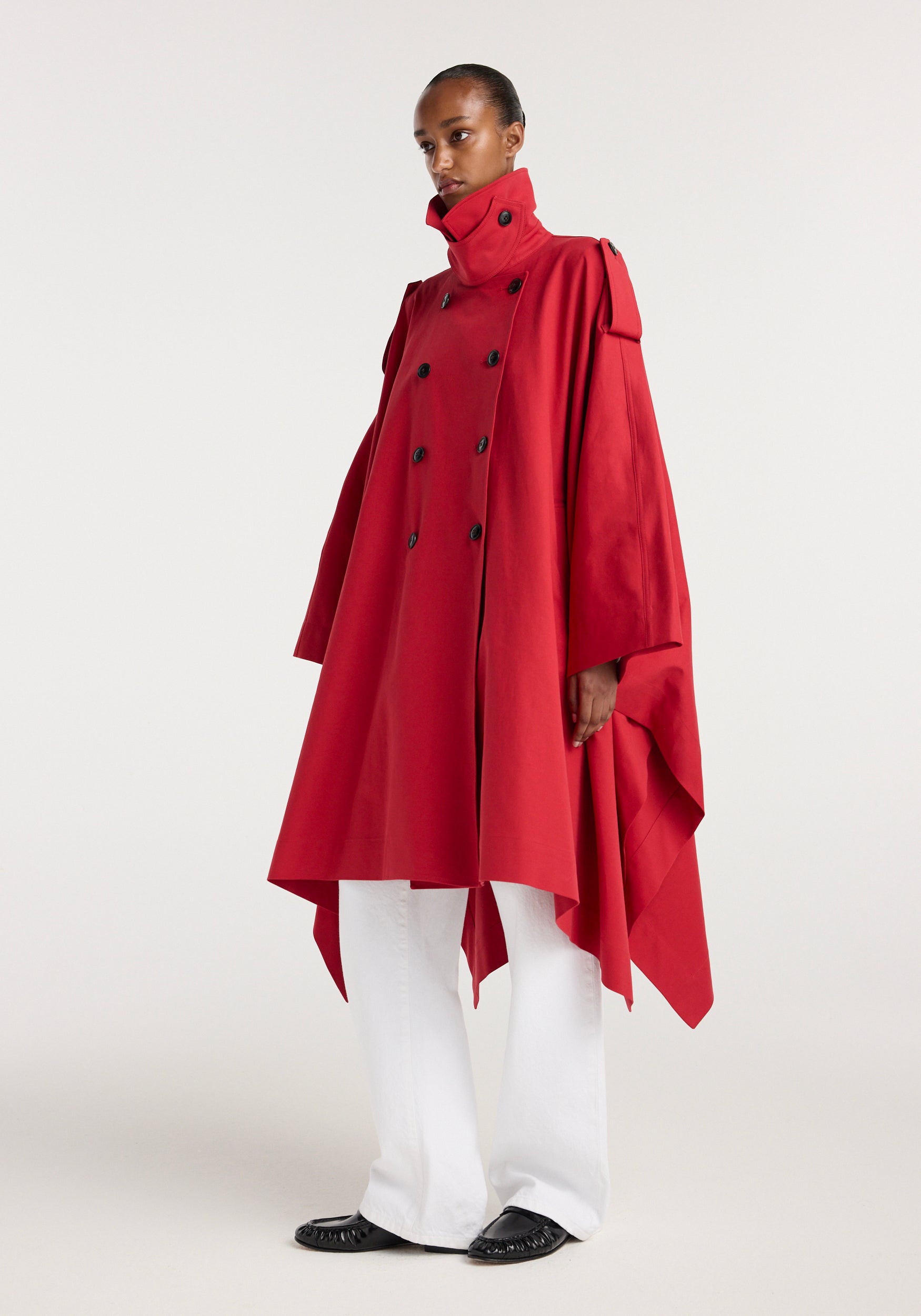 Model wearing a vibrant cherry red cape trenchcoat | cherry with a high collar, double-breasted front, and flowing asymmetrical hem paired with white pants and black shoes.