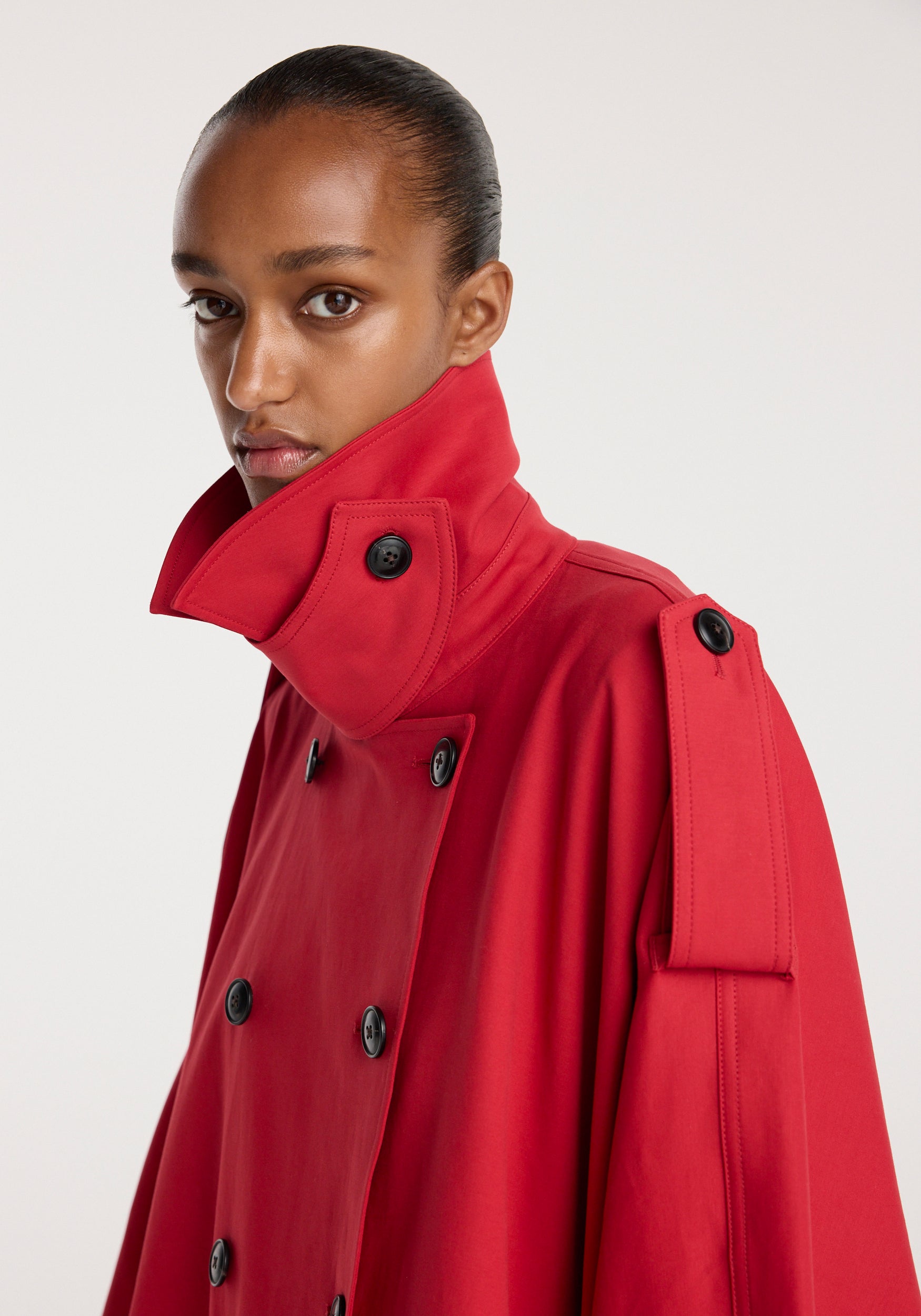 Close-up of a model wearing a vibrant red Cape trenchcoat | cherry with a high buttoned collar and black buttons, highlighting the structured shoulder detail against a plain background.