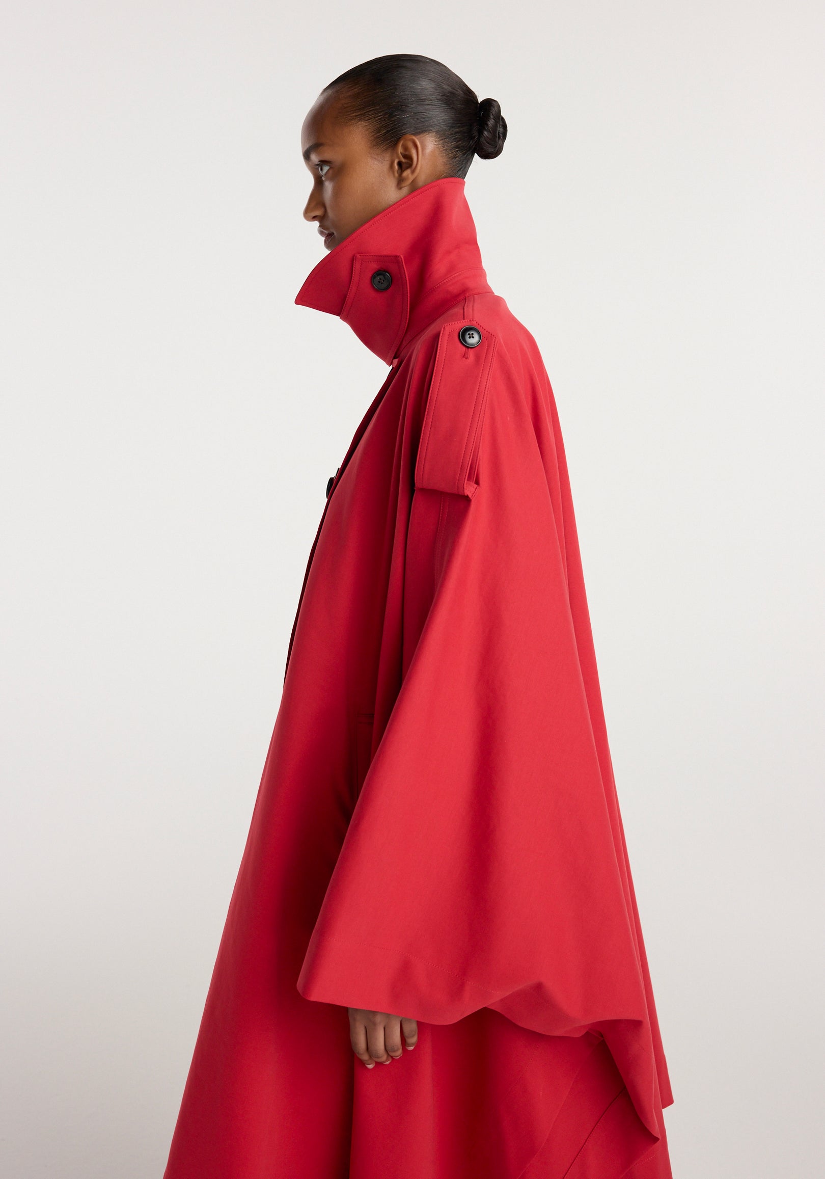 Side profile of a woman wearing a vibrant cherry red cape trenchcoat | cherry with high collar and buttoned shoulder details against a plain background.