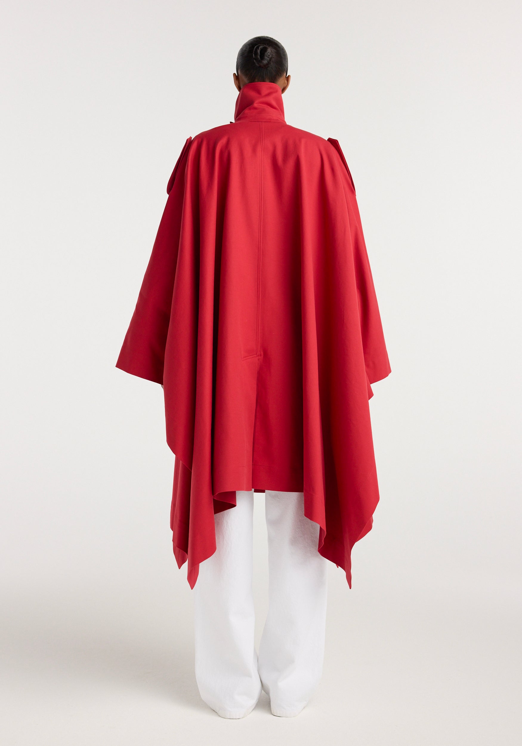 Back view of a person wearing a vibrant red Cape trenchcoat | cherry with a high collar and flowing cape-like silhouette, paired with white pants against a plain background.