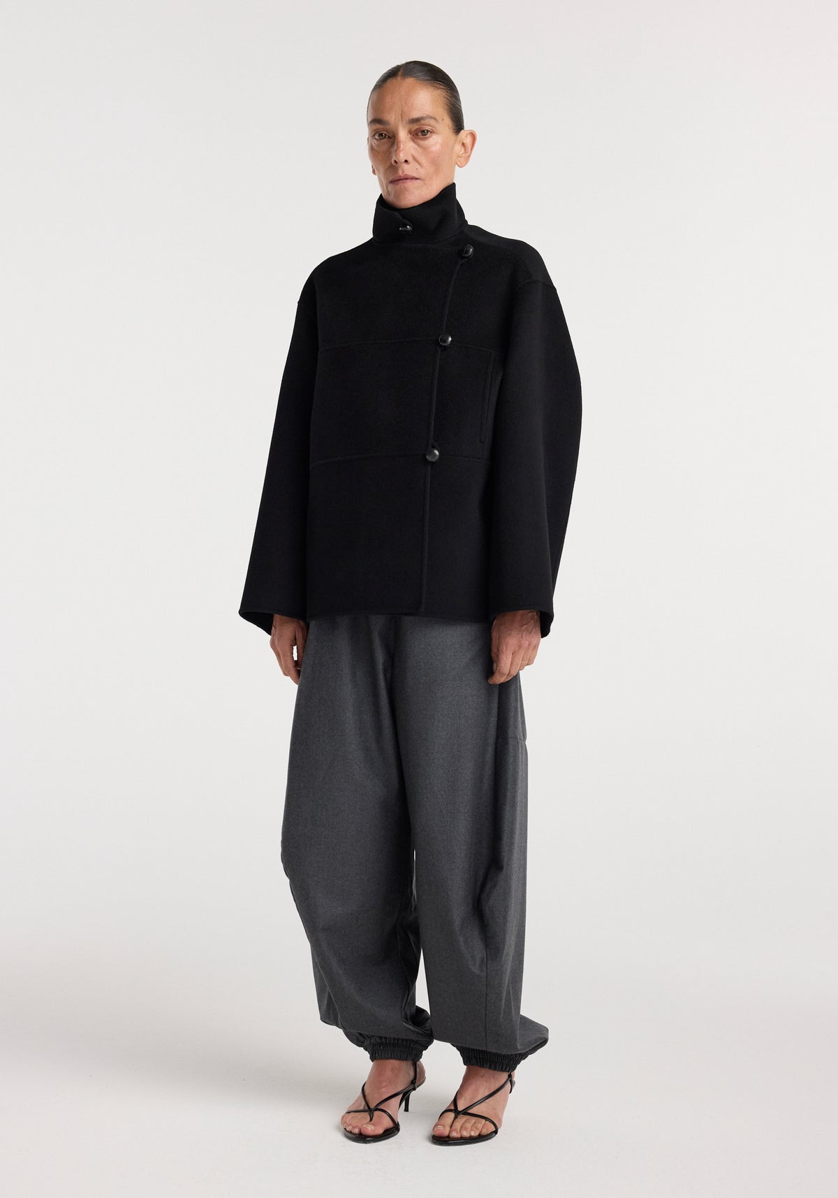 A model wearing a black Double-faced wool jacket | black with an asymmetrical button design, high collar, and loose fit paired with gray wide-leg pants and black strappy heels.