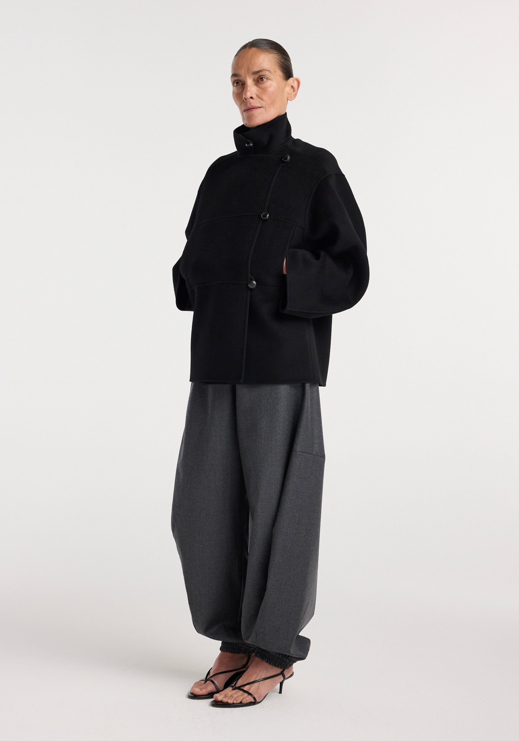 Woman wearing a black Double-faced wool jacket | black with an asymmetrical button closure, high collar, and wide sleeves, paired with loose gray trousers and black heeled sandals.