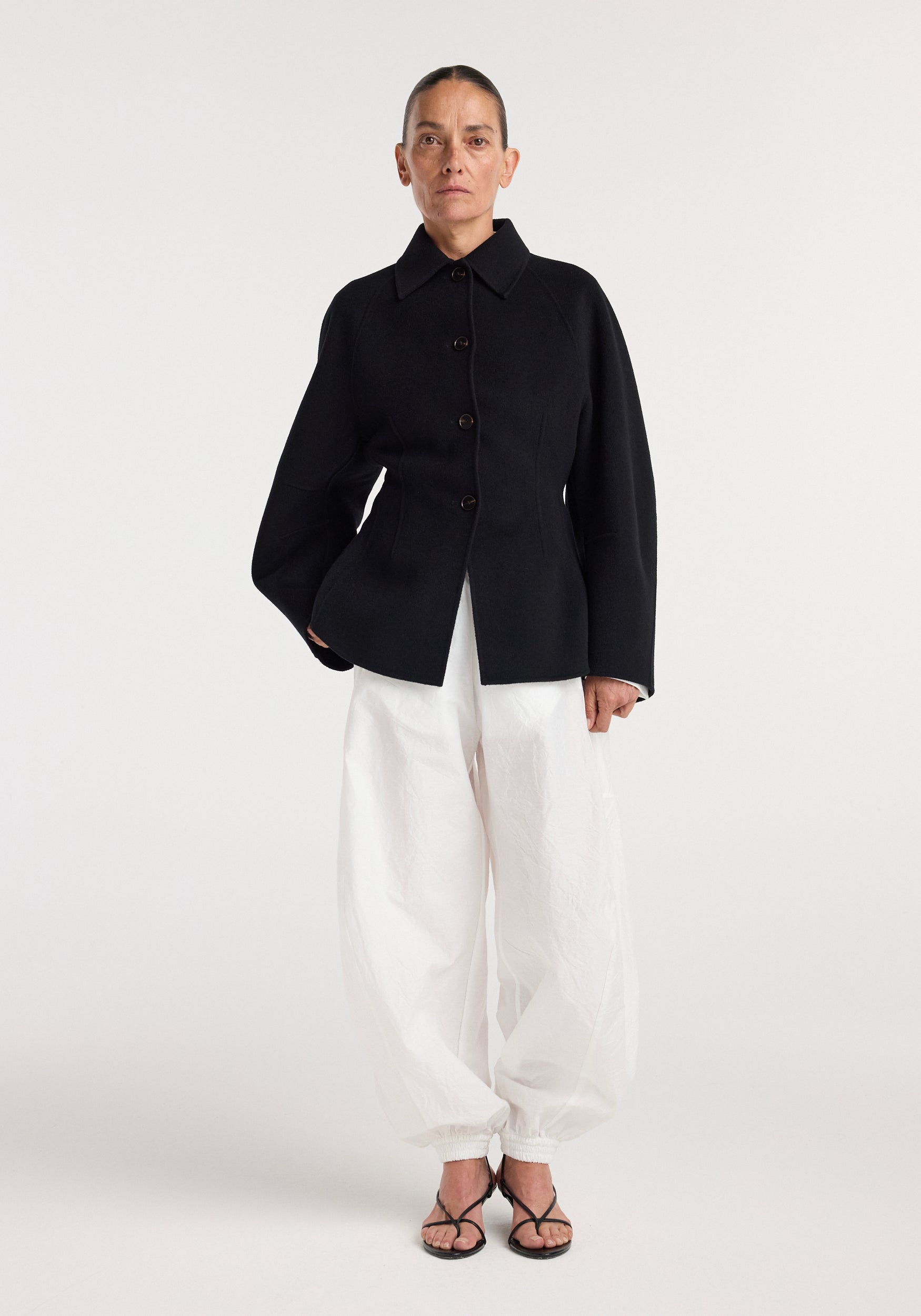 Woman wearing a black Double-faced sculptural jacket | black with a structured silhouette, paired with loose white pants and black strappy heels against a plain background.