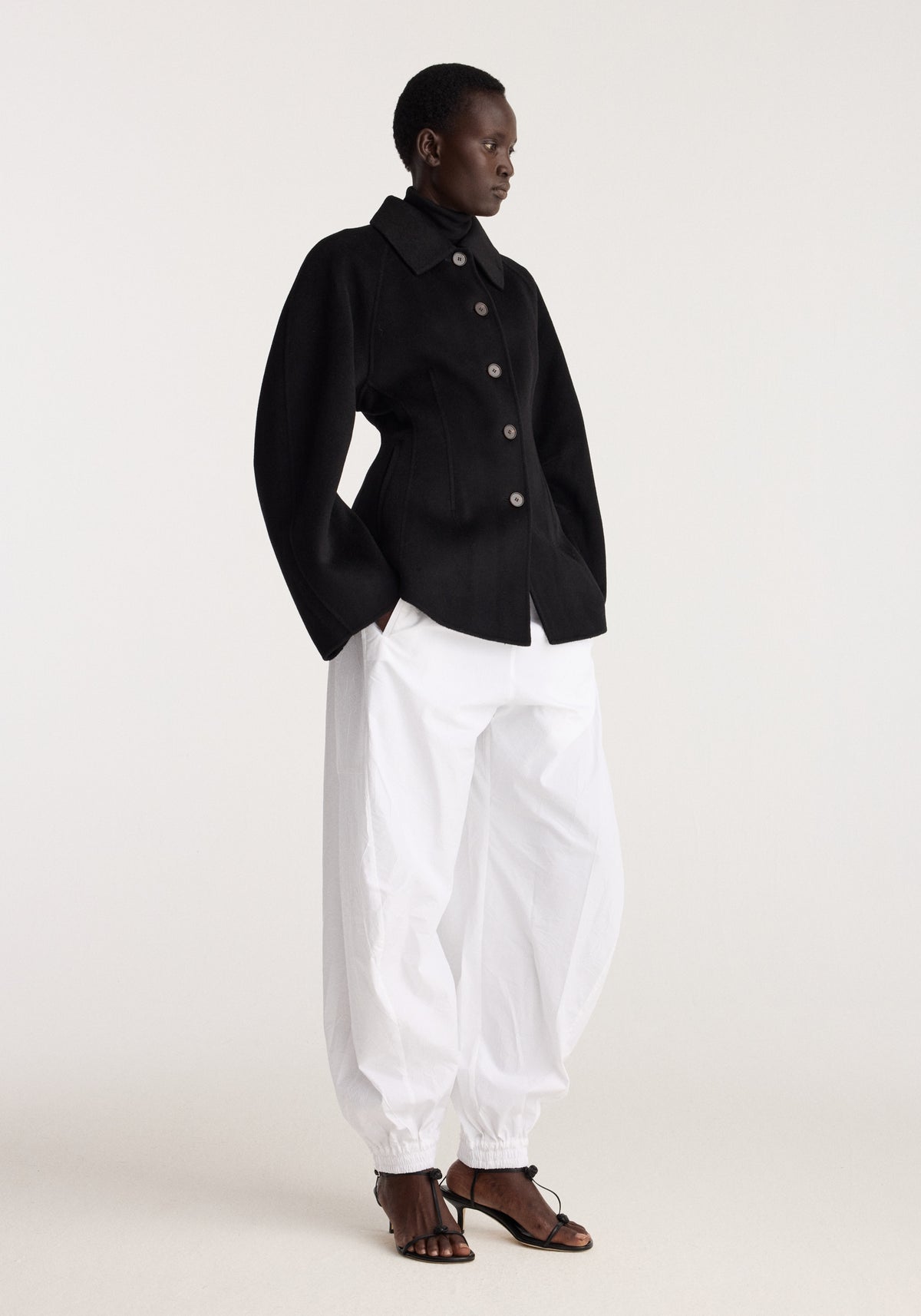 Double-faced sculptural jacket | black modeled with white trousers and flat strappy sandals in a minimalist studio setting