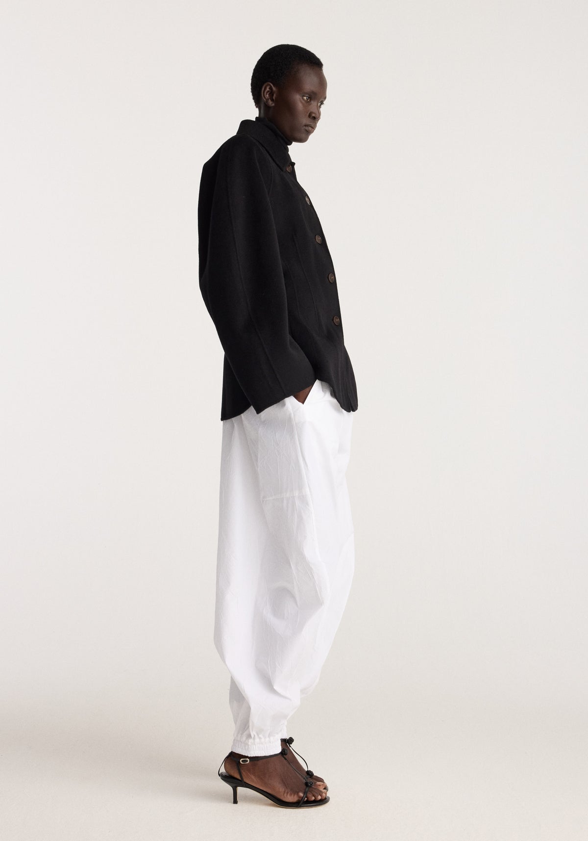 Double-faced sculptural jacket | black shown in side profile with white trousers and brown strappy sandals in a minimalist studio setting