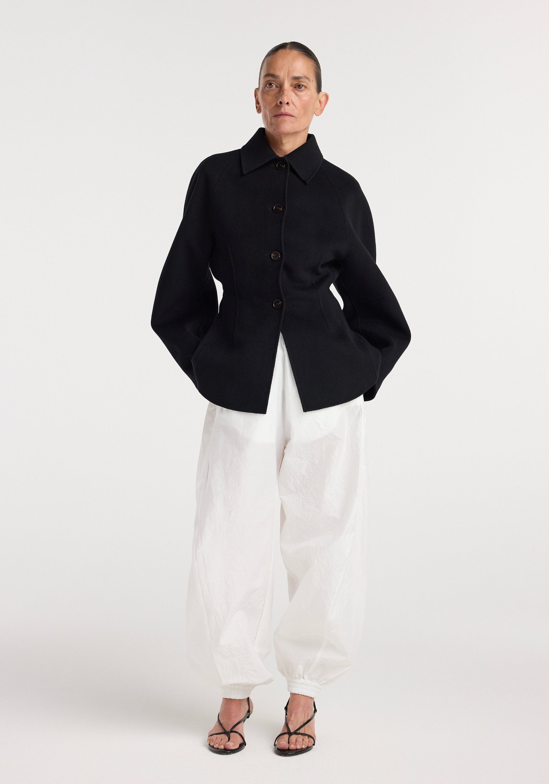 A model wearing a tailored black Double-faced sculptural jacket | black with structured sleeves and buttons, paired with loose white pants and black strappy sandals against a plain background.