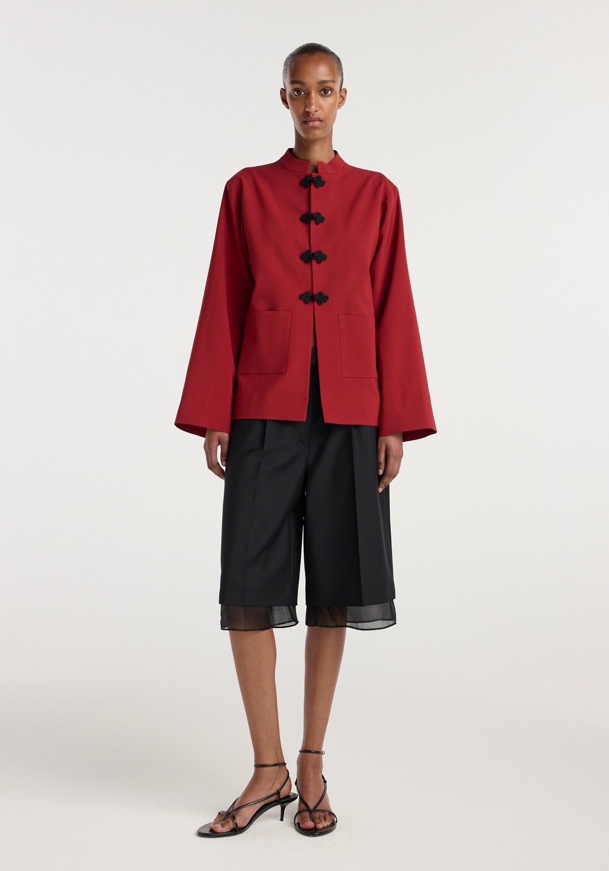 Model wearing a carmine Pankou closure jacket with wide sleeves and black frog button fastenings, paired with black wide-leg knee-length shorts and strappy black heels.