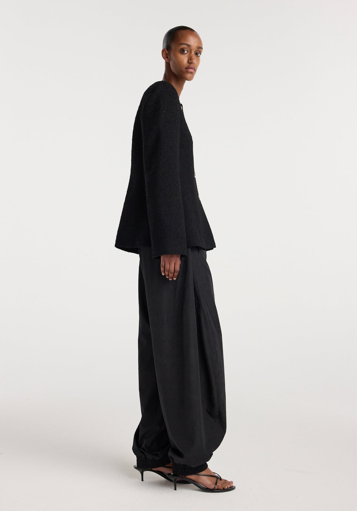 Model wearing a black Bouclé shaped blazer | black with long sleeves, paired with loose black pants and black open-toe heeled sandals in a minimalist studio setting.