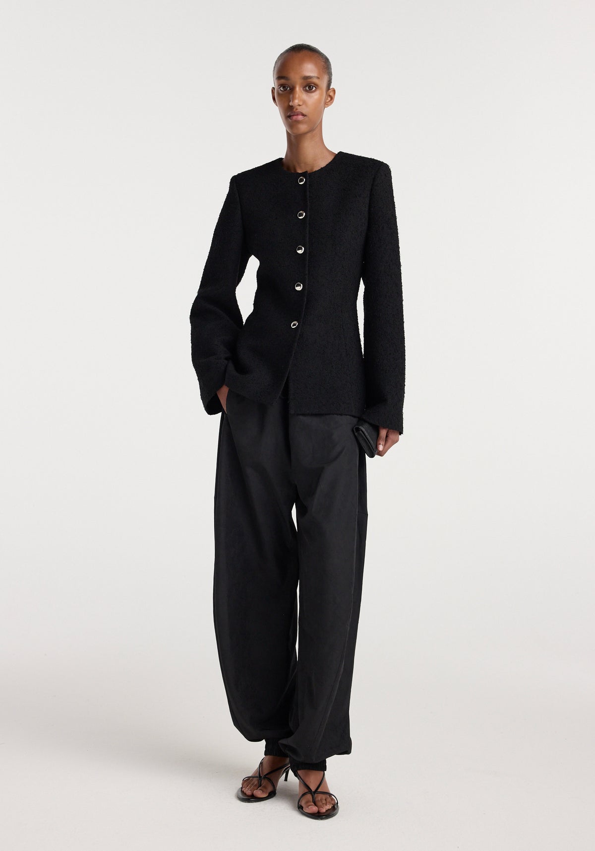 Woman wearing a black bouclé shaped blazer | black with long sleeves and front buttons, paired with loose black pants and black strappy sandals on a white background.