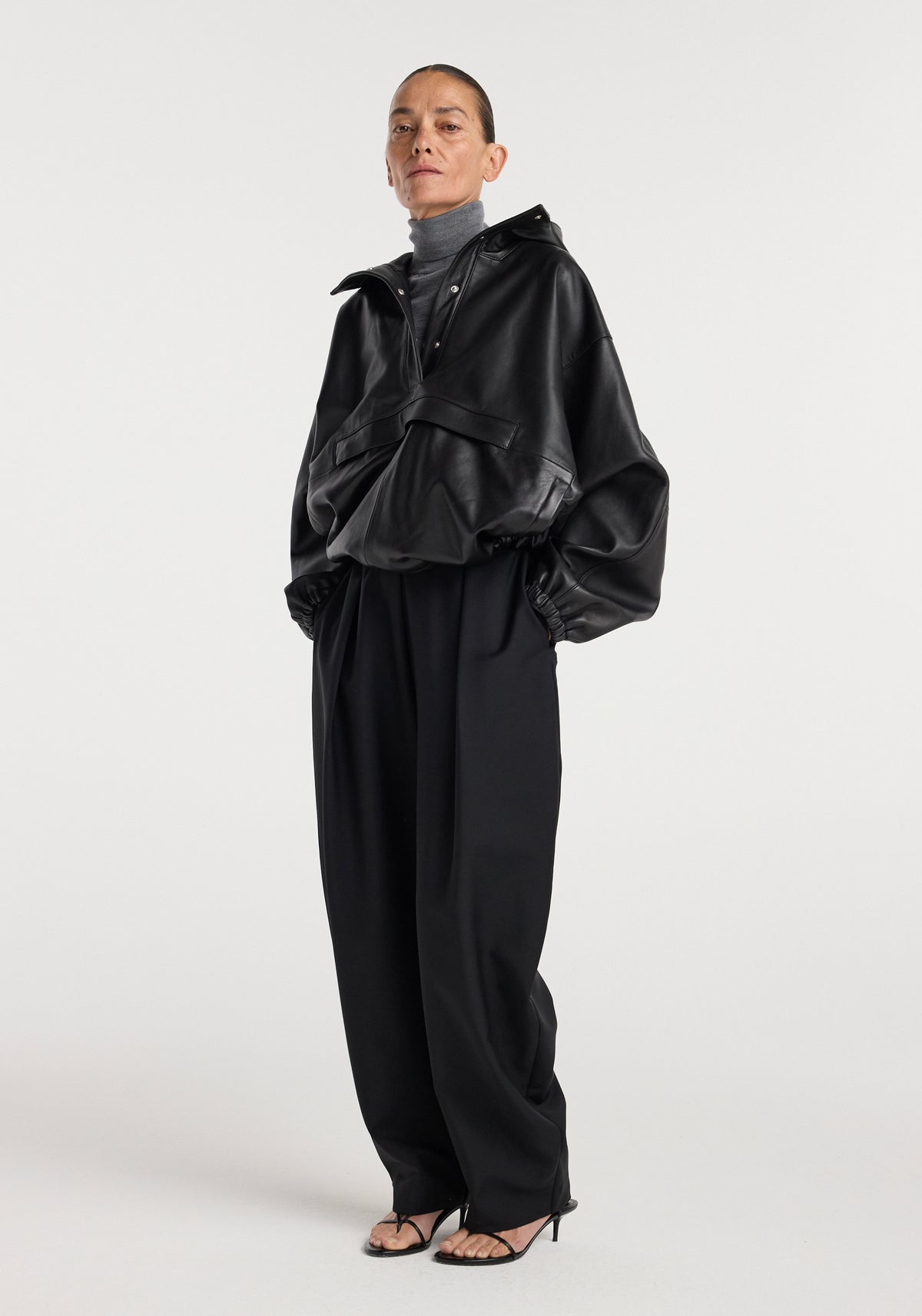 Model wearing a black Leather oversized anorak | black with long balloon sleeves, paired with black wide-leg pants, a gray turtleneck, and black open-toe heels, standing against a plain background.
