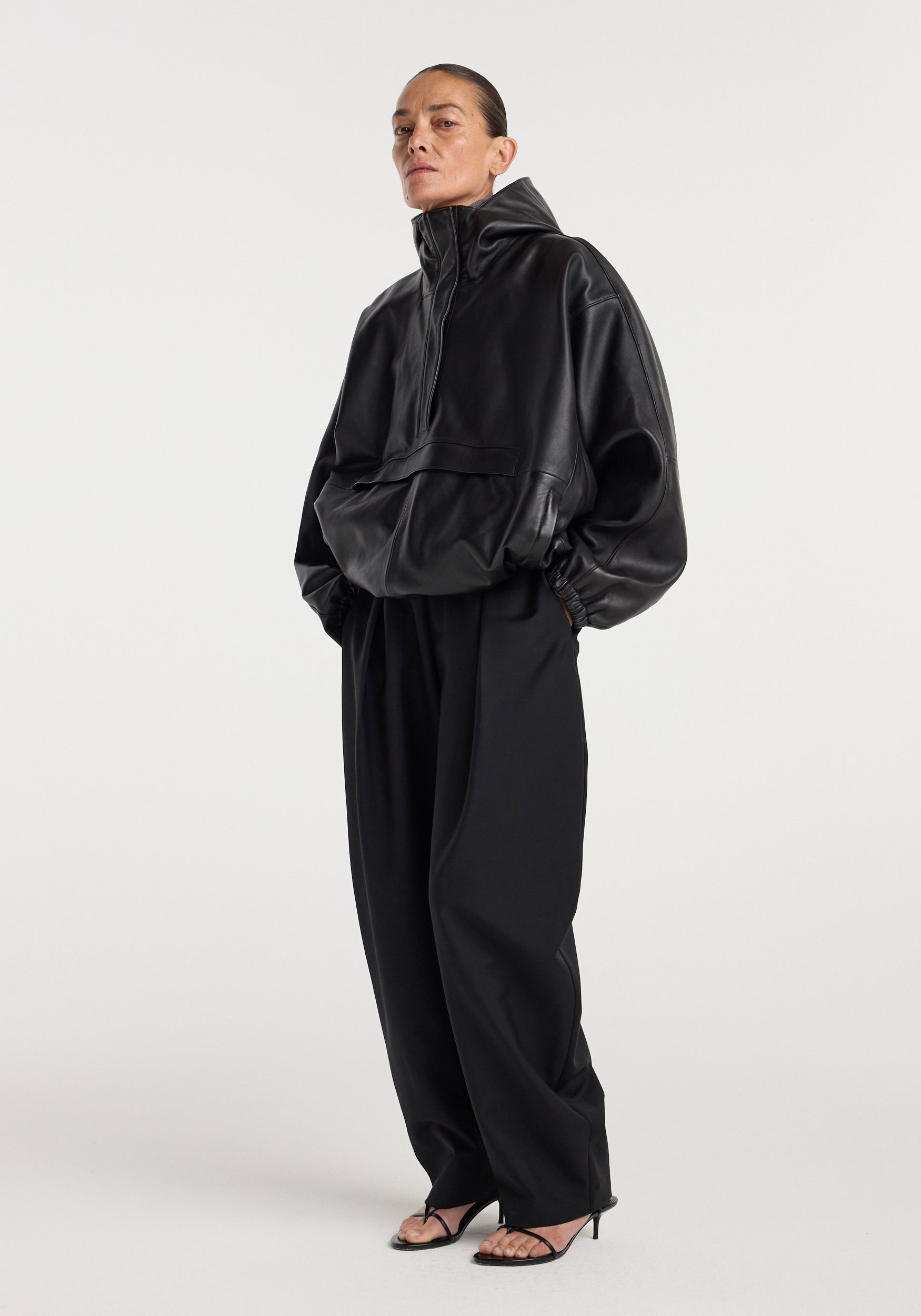 Woman wearing a black leather oversized anorak with a high collar and front pocket, paired with wide-leg black pants and black open-toe heels, standing against a plain background.