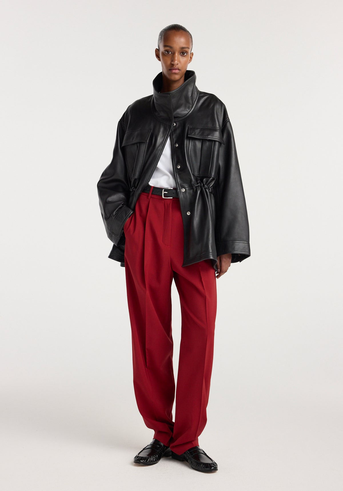 Model wearing a black sculptural leather jacket | black with oversized fit and high collar, paired with white top, red wide-leg trousers, and black shoes against a plain background.