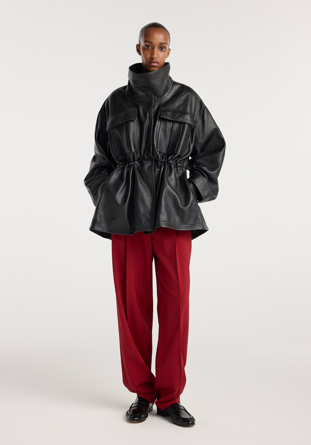 Model wearing a black sculptural leather jacket | black with a high collar and cinched waist, paired with wide-legged red trousers and black shoes against a plain background.