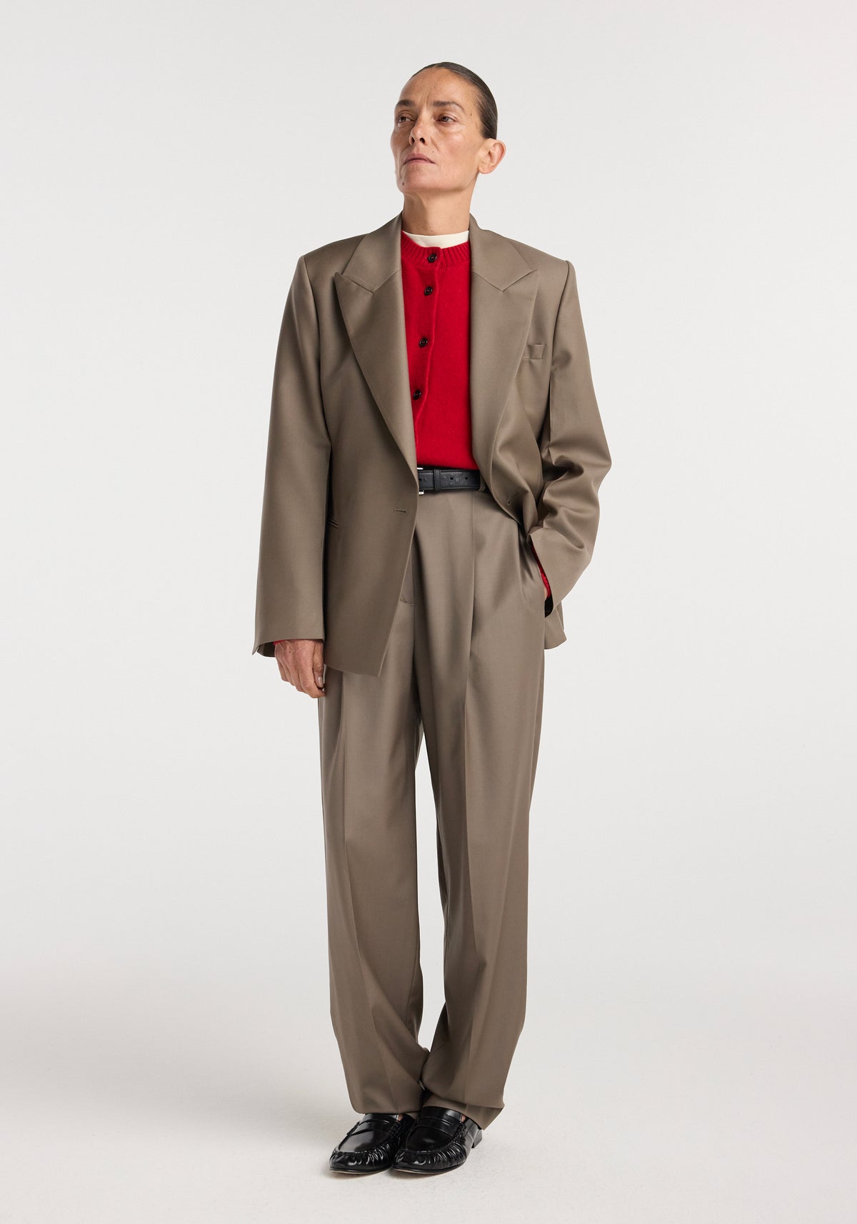 A model wearing an hourglass shaped blazer | desert brown paired with matching high-waisted trousers, a red buttoned top, black belt, and black shoes against a plain white background.