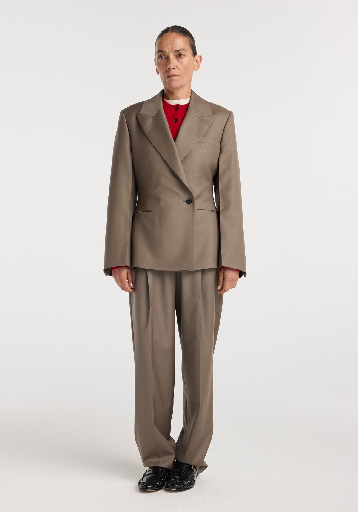 Woman wearing an hourglass shaped blazer | desert brown with matching wide-leg trousers and a red top underneath, standing against a plain white background.