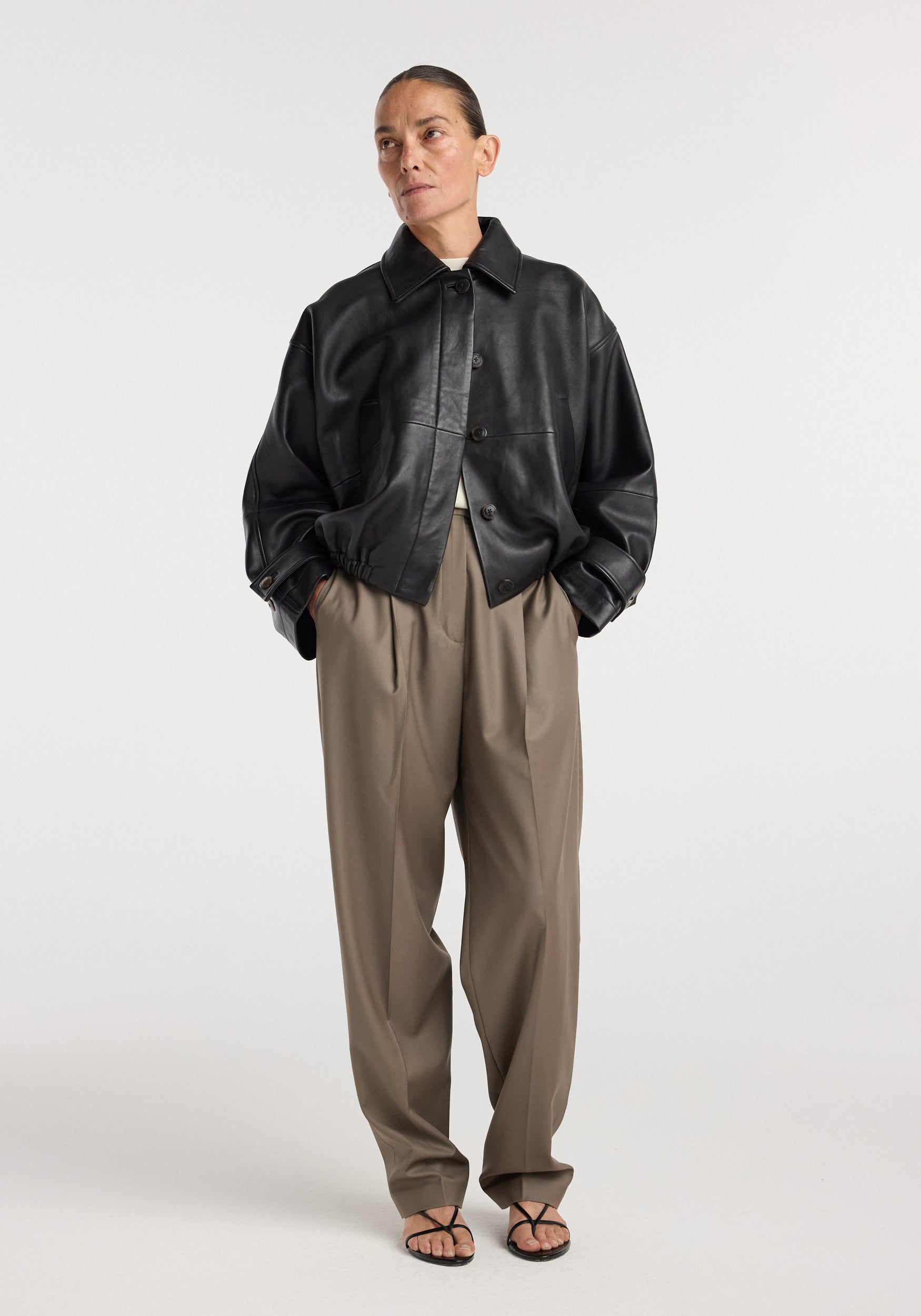 Woman wearing a black Leather blouson jacket | black with loose beige trousers and black strappy sandals standing against a plain white background.
