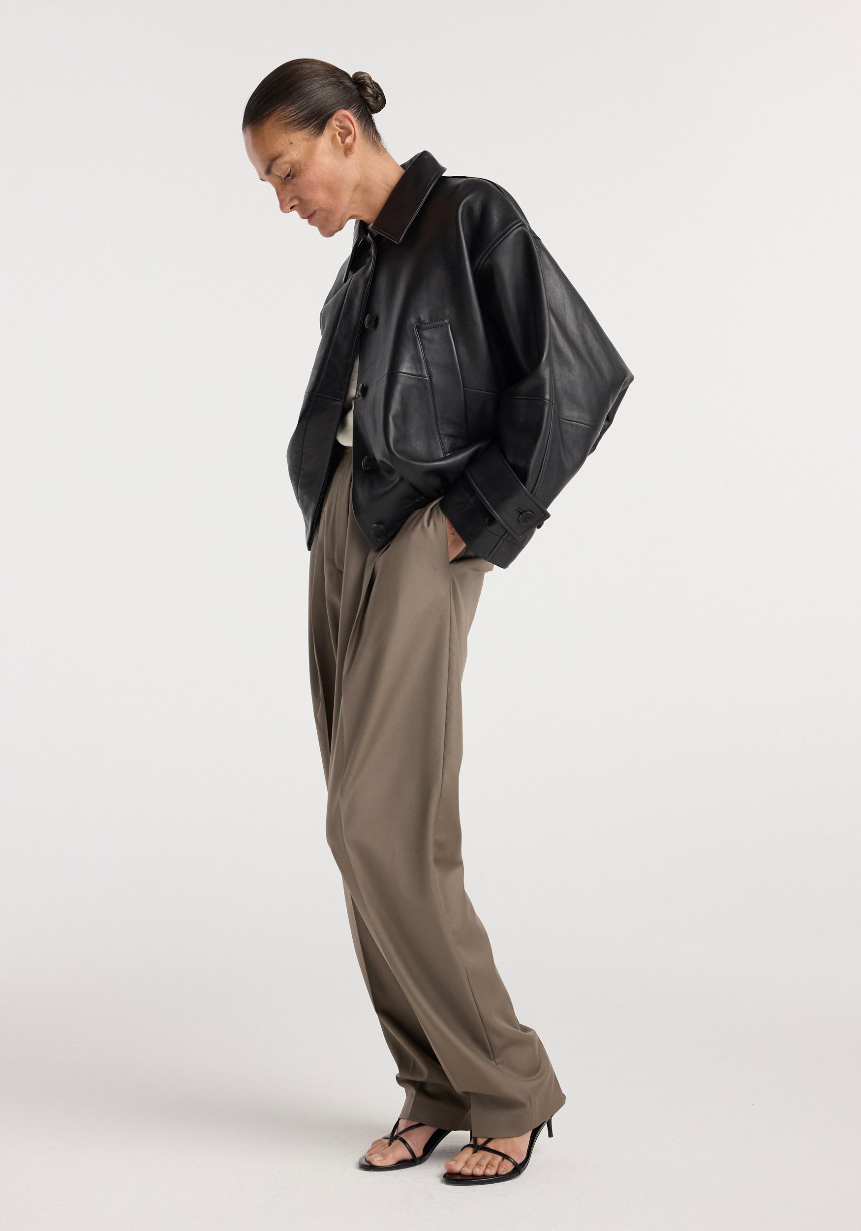 Woman wearing a black leather blouson jacket | black paired with loose brown trousers and black strappy heels, standing against a plain white background.