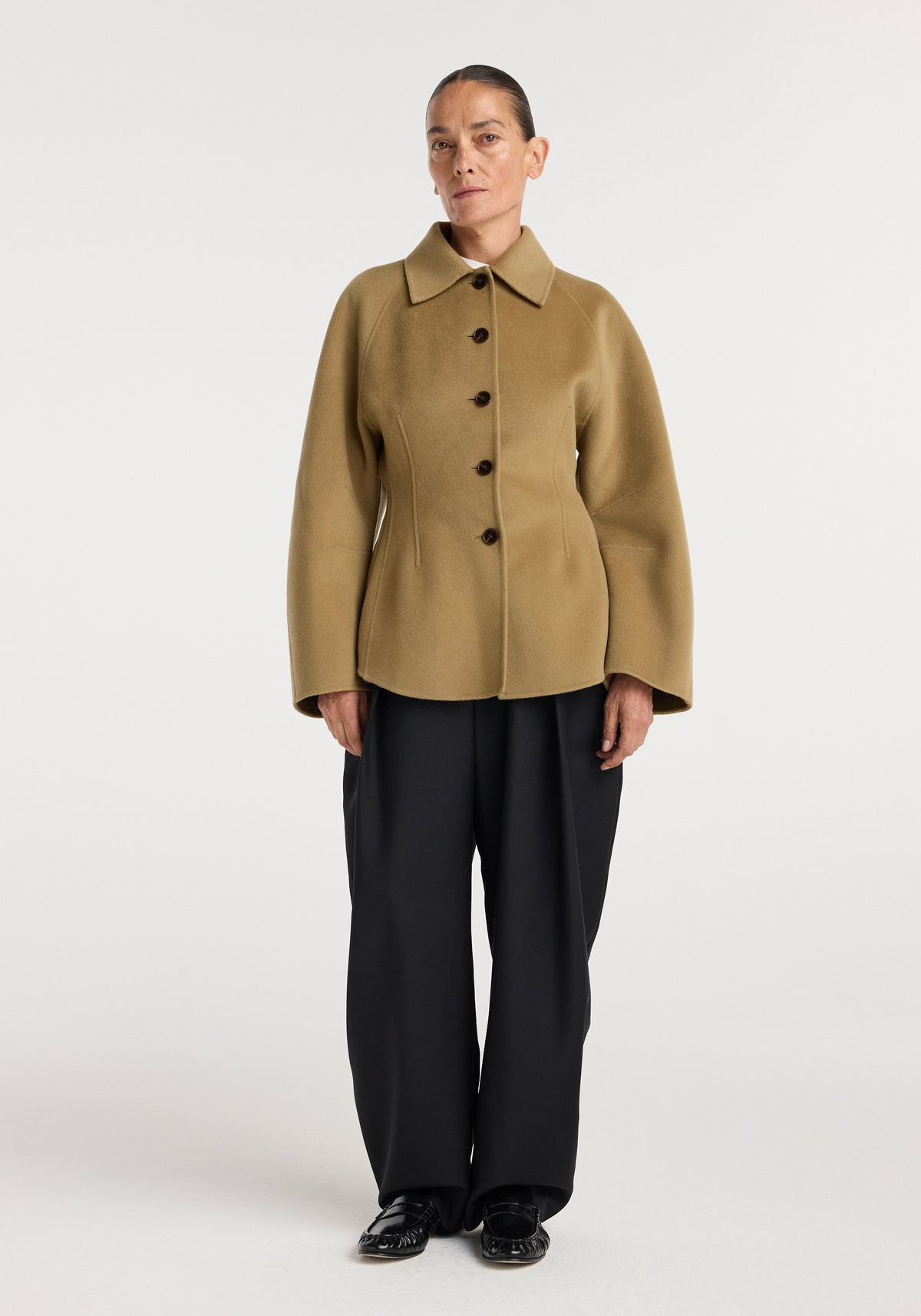 Woman wearing a camel color Double-faced sculptural jacket with a pointed collar and black buttons, paired with loose black pants and black shoes against a plain background.