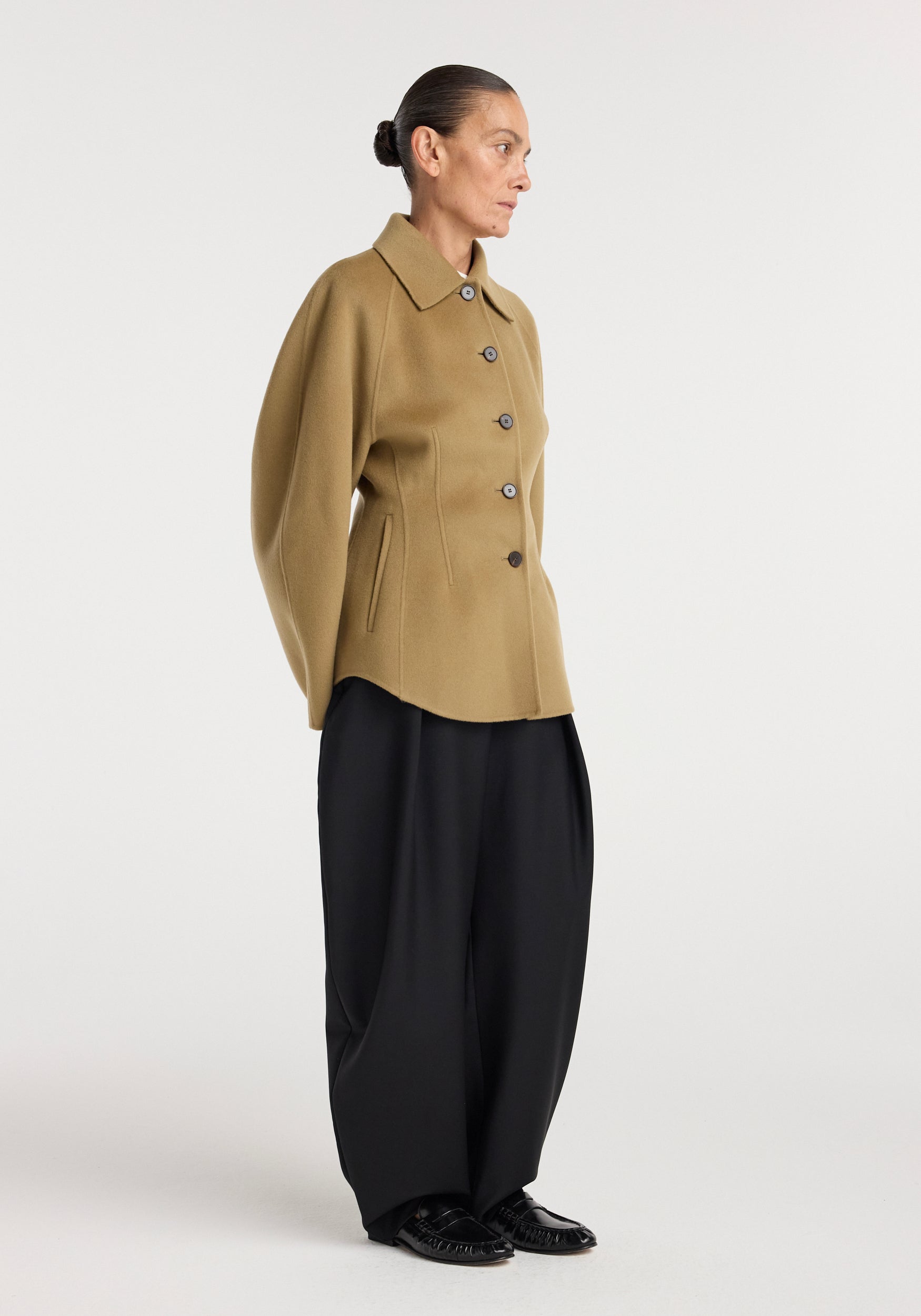 Woman modeling a camel-colored, double-faced sculptural jacket | camel with structured sleeves and front buttons, paired with loose black pants and black shoes against a plain background.