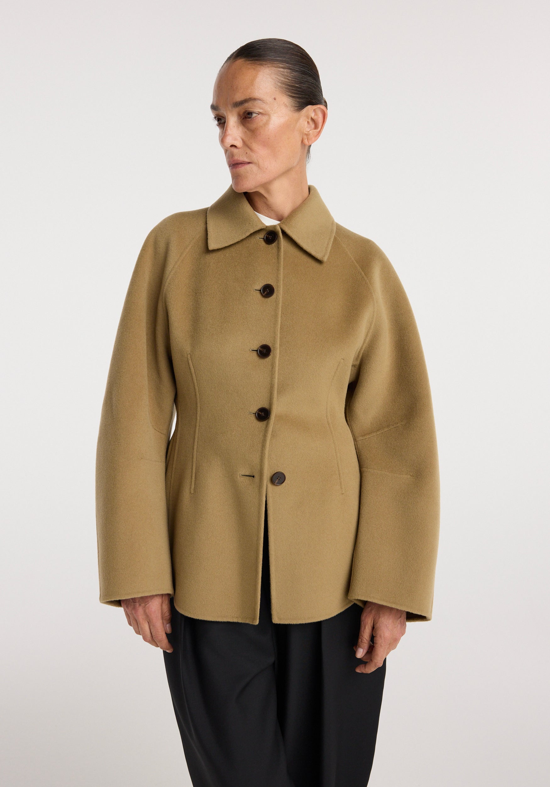 Woman wearing a camel-colored double-faced sculptural jacket with a wide collar and button closure, styled with black pants against a plain background.