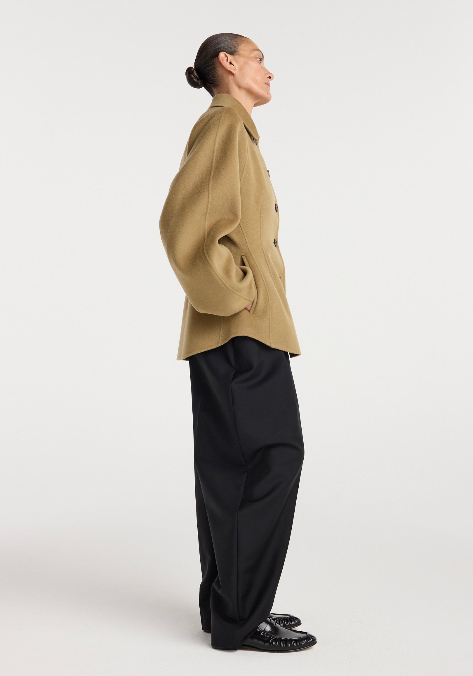 Side profile of a person wearing a camel-colored Double-faced sculptural jacket | camel with oversized sleeves and black wide-leg trousers against a plain white background.