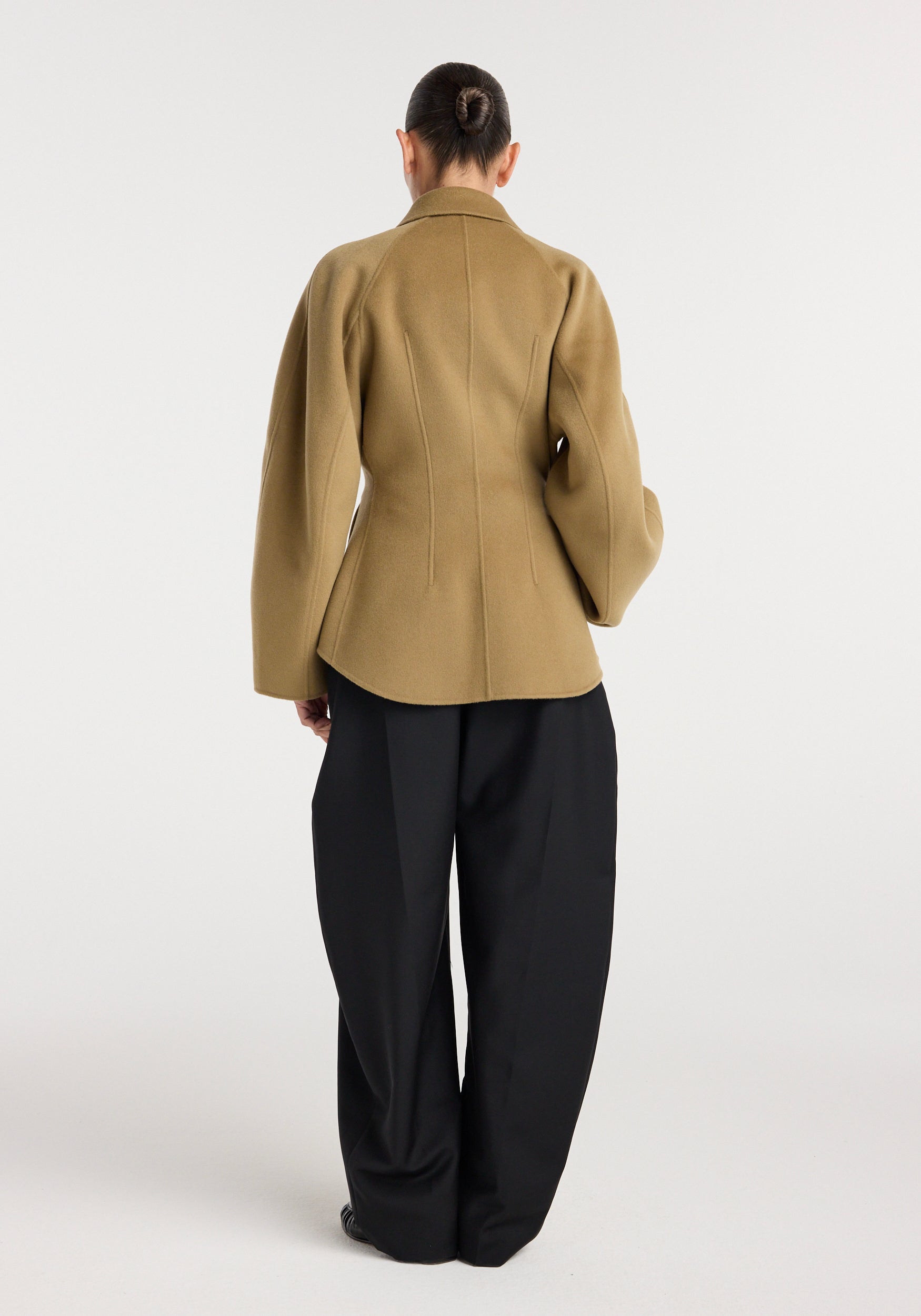 Back view of a person wearing a camel-colored double-faced sculptural jacket with unique tailored seams and voluminous sleeves, paired with loose black trousers against a plain background.