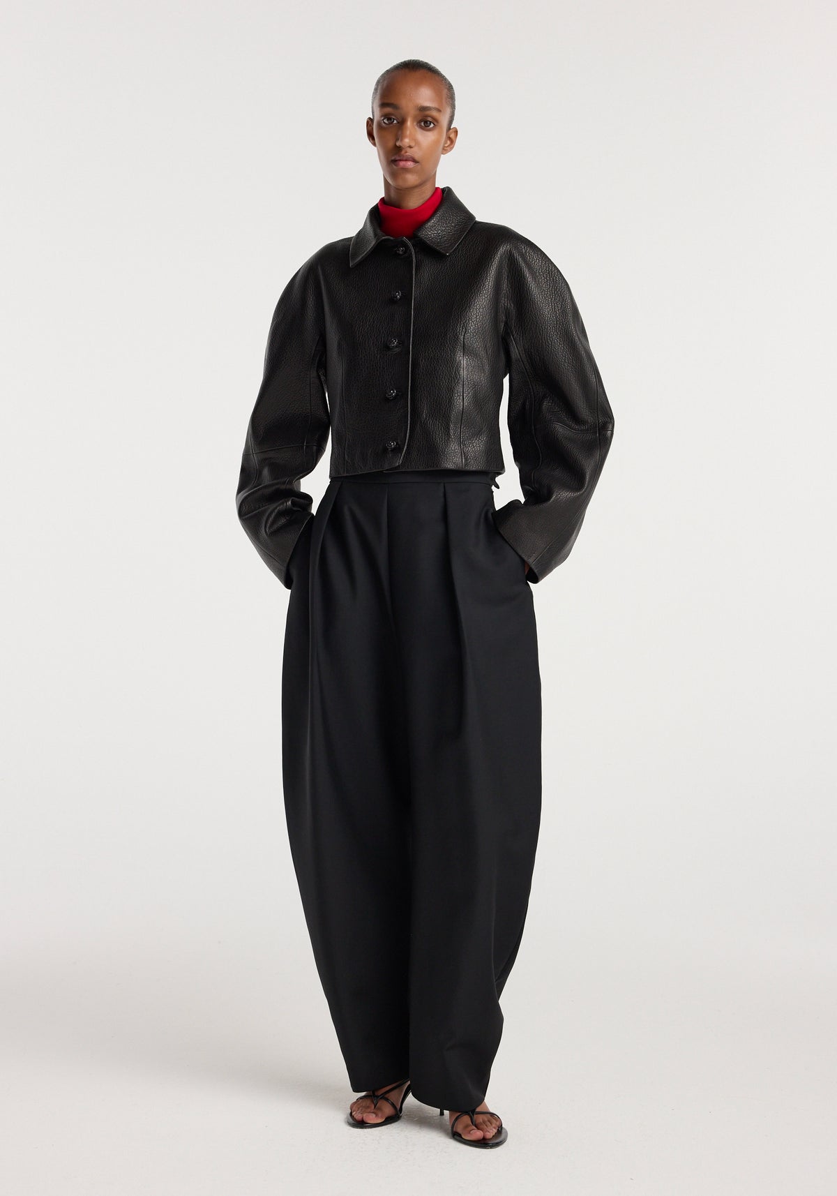 Model wearing a black rounded grain leather cropped jacket | black with long puffed sleeves, button closure, and a classic collar, paired with high-waisted black wide-leg pants and a red turtleneck underneath.