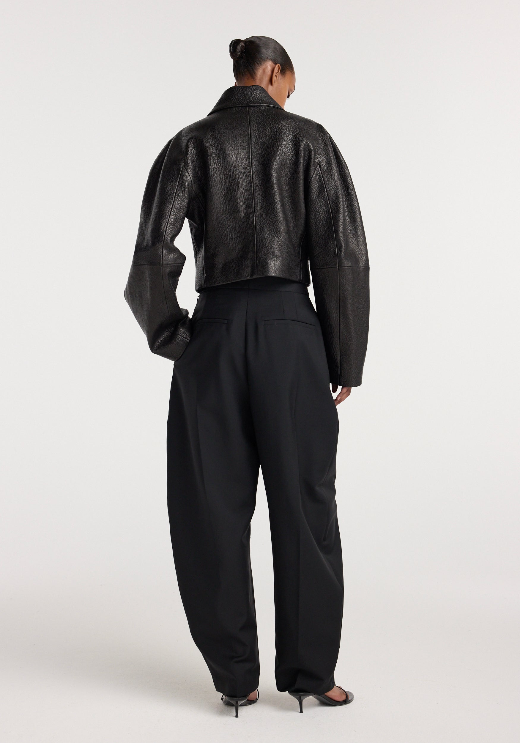 Woman wearing a black Rounded grain leather cropped jacket | black, paired with loose black high-waisted pants and black heels, shown from the back against a plain background.