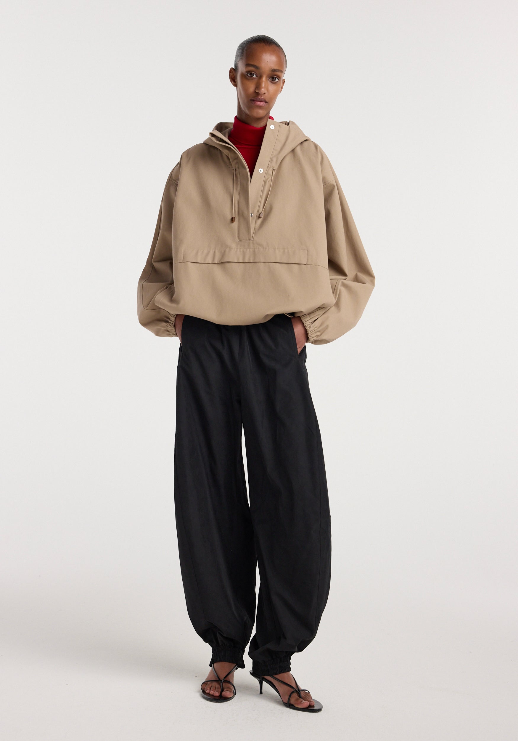 Model wearing a beige Hooded anorak with elastic cuffs and a front pocket, paired with loose black pants and strappy black heeled sandals against a plain background.