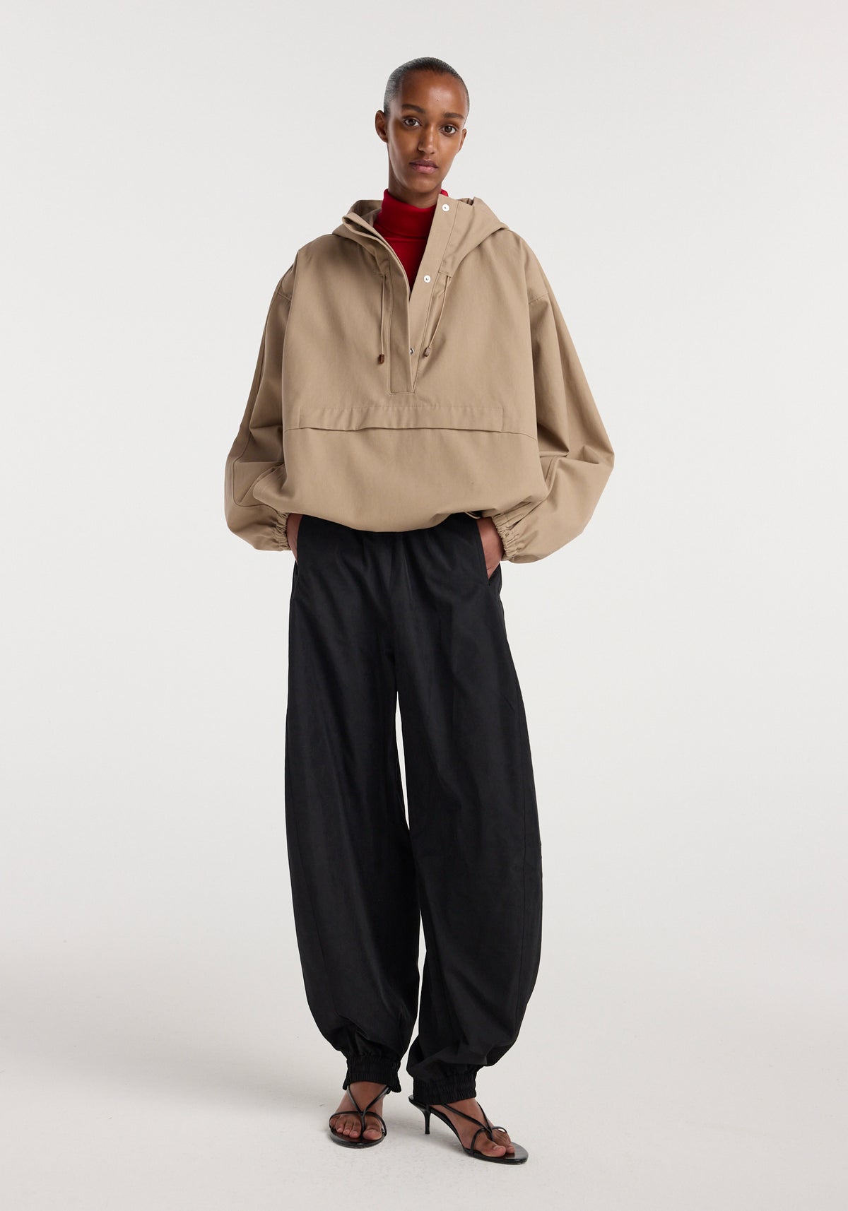 Model wearing a beige Hooded anorak with elastic cuffs and a front pocket, paired with loose black pants and strappy black heeled sandals against a plain background.